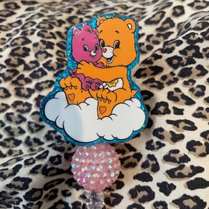 May include: A badge reel featuring two Care Bears hugging on a cloud. The bears are orange and pink, with a blue glitter border. The reel has a pink, glittery ball and a clear plastic clip.