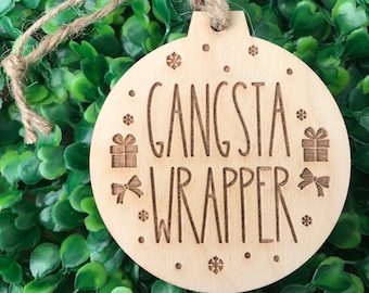 May include: A wooden Christmas ornament shaped like a circle with the text "Gangsta Wrapper" and snowflakes and gift boxes around the text.