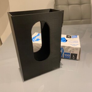 May include: A black 3D printed dispenser for holding a box of disposable gloves. The dispenser has a slot for the box to slide into and a cutout for the gloves to be dispensed from. The box of gloves is visible in the background, with the text "INTCO" and "100 GLOVES" printed on it.