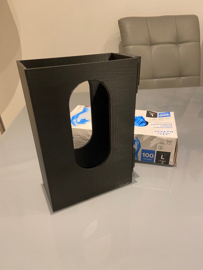 May include: A black 3D printed dispenser for holding a box of disposable gloves. The dispenser has a slot for the box to slide into and a cutout for the gloves to be dispensed from. The box of gloves is visible in the background, with the text "INTCO" and "100 GLOVES" printed on it.