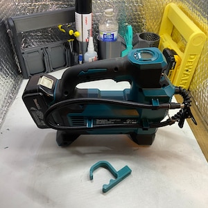 Puede incluir: Un compresor de aire inalámbrico azul turquesa con una manguera negra y un manómetro digital. El compresor tiene un mango negro y una batería negra. El compresor está sobre una superficie blanca.