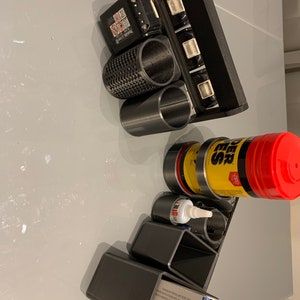 May include: A black 3D printed organizer with multiple compartments for holding tools and supplies. The organizer has a handle on the top and is filled with various items, including a black battery, a yellow and black bottle of glue, a red and black bottle of cleaner, and a black and white box.