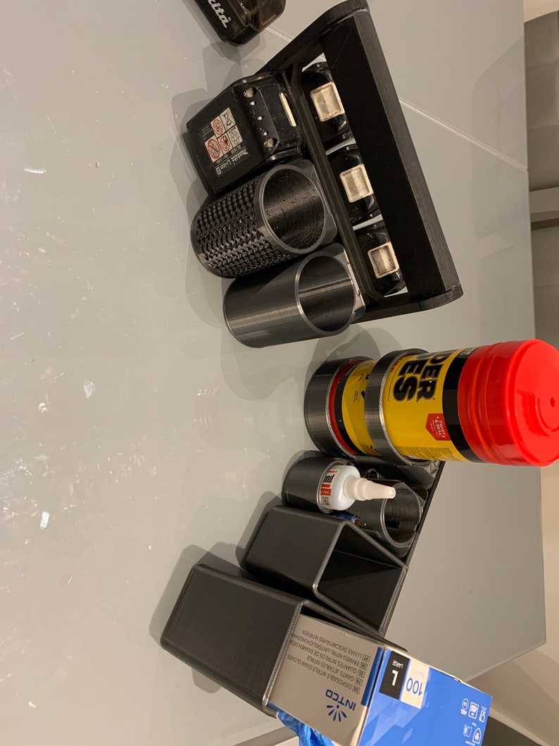 May include: A black 3D printed organizer with multiple compartments for holding tools and supplies. The organizer has a handle on the top and is filled with various items, including a black battery, a yellow and black bottle of glue, a red and black bottle of cleaner, and a black and white box.