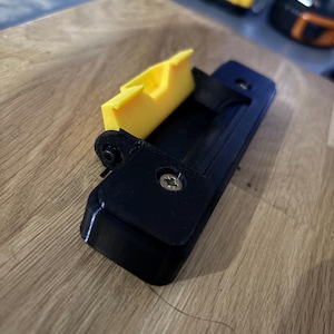May include: A black and yellow tool component, possibly for a larger device. The component has a rectangular black base with a yellow, angled piece attached. A screw secures the yellow piece. The item rests on a wooden surface.
