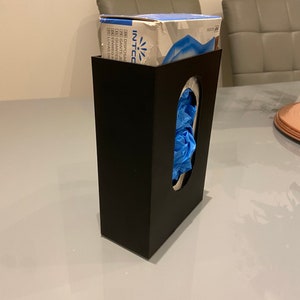 May include: A black plastic dispenser for disposable gloves. The dispenser has a slot for the gloves to be pulled out of. A box of blue disposable gloves is sitting on top of the dispenser. The box has the text "INTCO" on it.