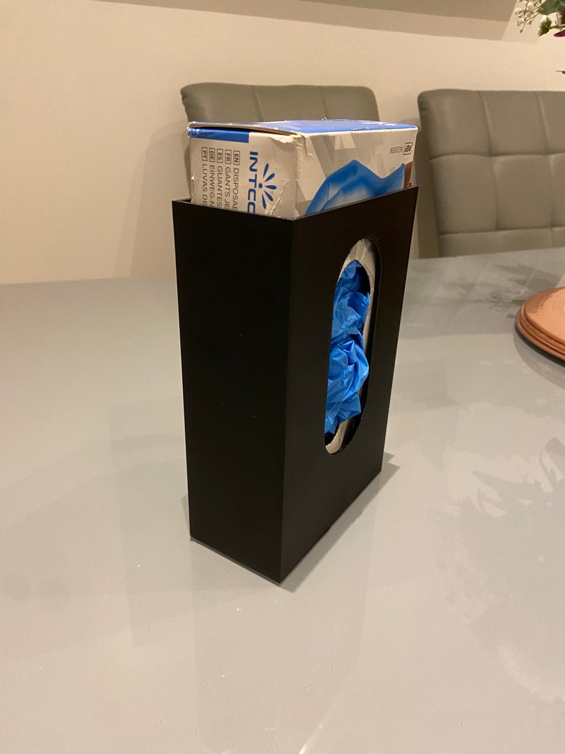 May include: A black plastic dispenser for disposable gloves. The dispenser has a slot for the gloves to be pulled out of. A box of blue disposable gloves is sitting on top of the dispenser. The box has the text "INTCO" on it.