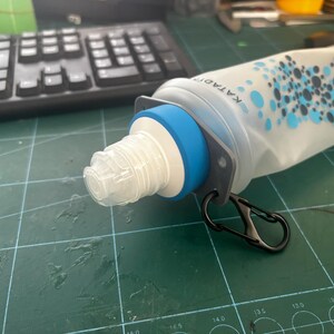 May include: A water bottle with a white and blue design. The bottle has a blue ring around the neck and a black carabiner clip. The bottle is lying on a green surface near a black keyboard.