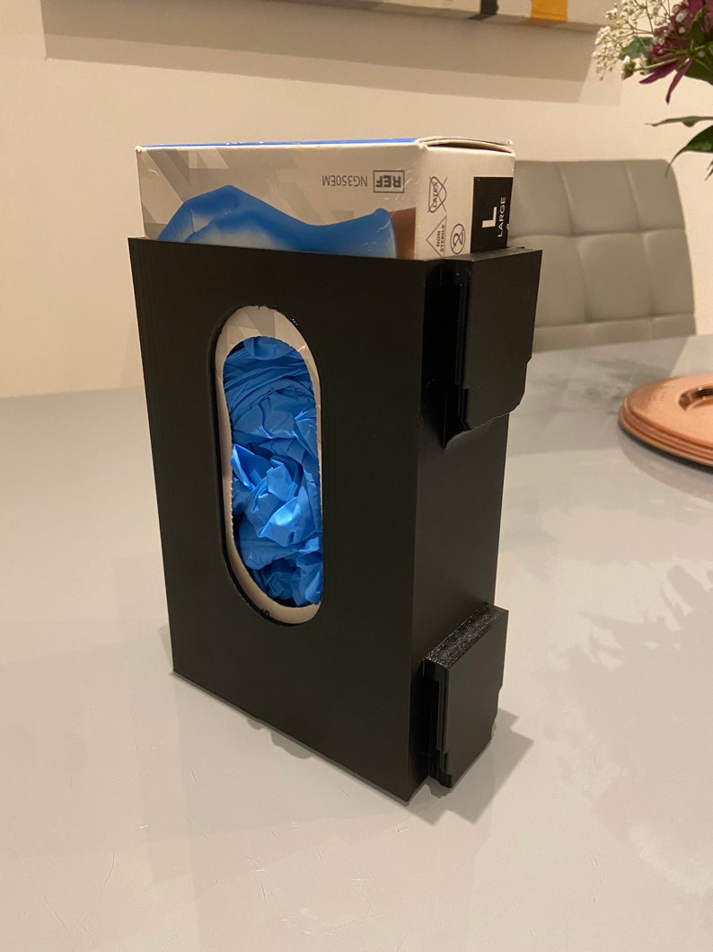 May include: Black plastic dispenser for disposable gloves with a box of blue nitrile gloves on top. The dispenser has a slot for dispensing gloves and a label that says "REF. NGE503M".