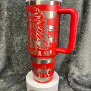 May include: Red stainless steel travel mug with a red lid and handle. The mug features a silver eagle design with the words "The Few, The Proud, The Fearful Men" engraved on it.