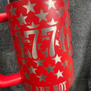 May include: Red stainless steel tumbler with a silver design of stars and the number 1776. The text "We Are Not Done" is printed on the bottom of the tumbler.