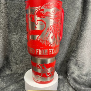 May include: Red stainless steel tumbler with a silver eagle and American flag design. The tumbler has a red lid and handle. The text "Unleashed From Fear" is engraved on the tumbler.