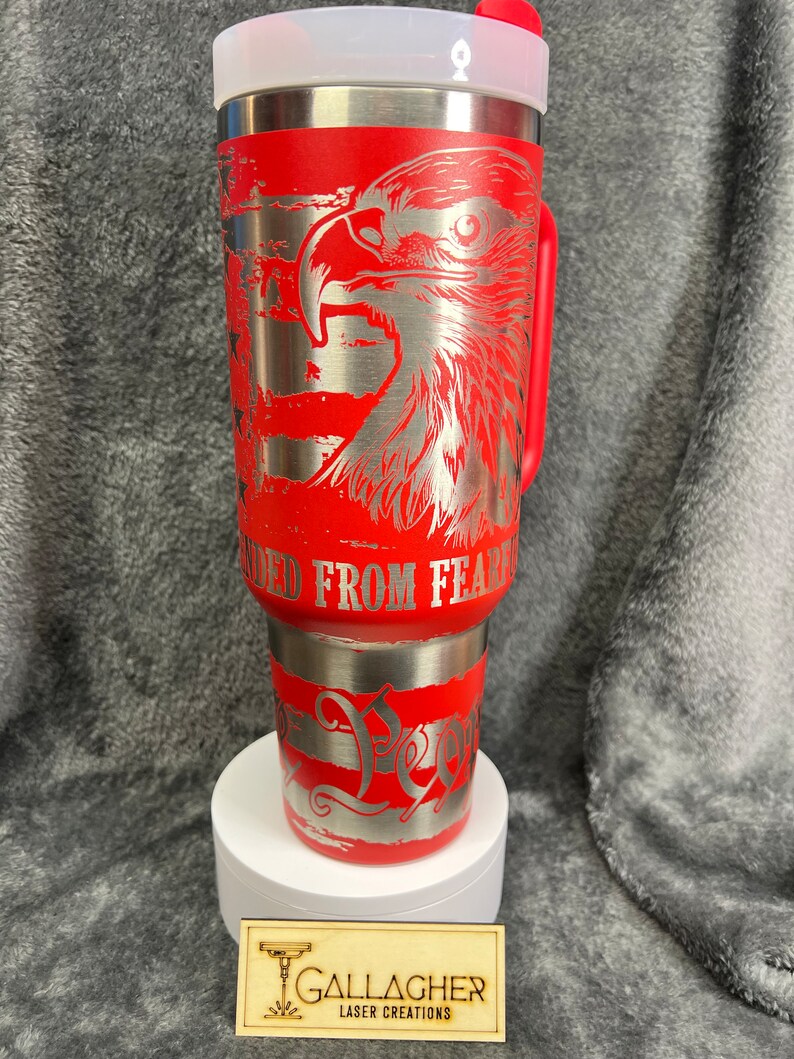 May include: Red stainless steel tumbler with a silver eagle and American flag design. The tumbler has a red lid and handle. The text "Unleashed From Fear" is engraved on the tumbler.