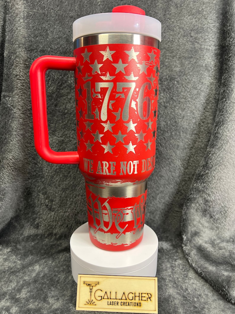 May include: Red stainless steel travel mug with a red handle and a white lid. The mug features a silver design of stars and the number 1776. The text "We are not done yet" is also on the mug.