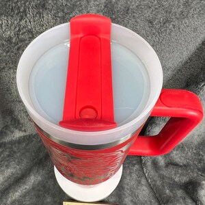 May include: Red travel mug with a white lid and a red silicone handle. The mug has a stainless steel exterior and a red and white design. The mug is sitting on a white coaster with the text "Gallagher Laser Creations" on it.