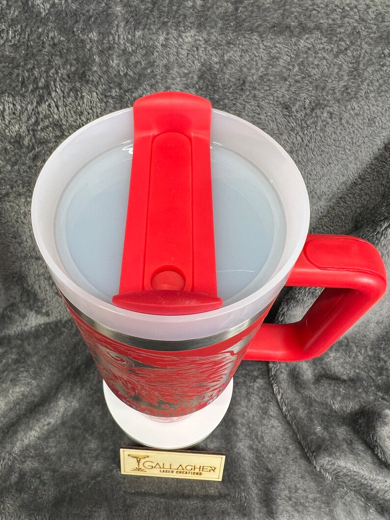 May include: Red travel mug with a white lid and a red silicone handle. The mug has a stainless steel exterior and a red and white design. The mug is sitting on a white coaster with the text "Gallagher Laser Creations" on it.