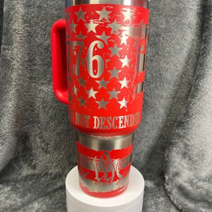 May include: Red stainless steel travel mug with a white lid and a red handle. The mug features a distressed American flag design with silver stars and the number "1776" on the front. The text "We Not Descended" is on the bottom of the mug.