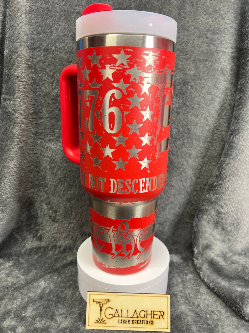 May include: Red stainless steel travel mug with a white lid and a red handle. The mug features a distressed American flag design with silver stars and the number "1776" on the front. The text "We Not Descended" is on the bottom of the mug.
