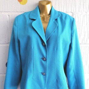 May include: A turquoise blazer with a notched lapel and three decorative buttons. The jacket is displayed on a mannequin. The background is a white brick wall with abstract art. The blazer has a tailored fit.