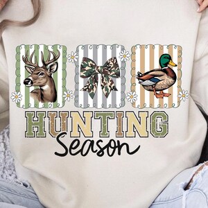 May include: A cream sweatshirt with three framed images: a deer, a camouflage bow, and a duck. The words "HUNTING Season" are printed below in green and brown, with a black script font below.