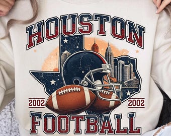 以下が含まれることがあります： クリーム色のスウェットシャツ。赤と青の文字で「HOUSTON FOOTBALL」とプリントされたグラフィック。デザインには、フットボール、ヘルメット、テキサスの輪郭、都市景観が含まれています。「2002」の年号がフットボールの両側に印刷されています。