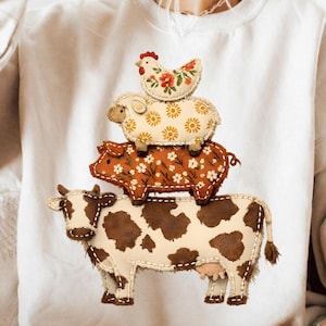 Könnte beinhalten: Ein cremefarbenes Sweatshirt mit einem Design von gestapelten Hoftieren: einer Kuh, einem Schwein, einem Schaf und einem Huhn. Die Kuh ist braun und weiß, das Schwein ist orange mit weißen Blumenmustern, das Schaf ist beige mit gelben Blumenmustern und das Huhn ist rot und floral.