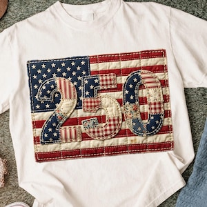 May include: White t-shirt with a patriotic design. The design features a patchwork American flag with the numbers "250" stitched on top. The flag has red and white stripes, blue stars, and the numbers are in a mix of patterns.