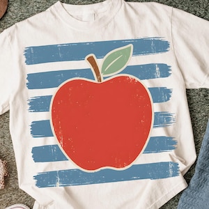 Puede incluir: Camiseta blanca con un gran gráfico de manzana roja, hoja verde y tallo marrón. La manzana se encuentra sobre trazos horizontales azules, creando un aspecto envejecido y vintage. Una prenda casual para el día a día.