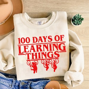 May include: A cream-coloured sweatshirt featuring the red text "100 DAYS OF LEARNING THINGS". Below the text are three red bicycle figures. The sweatshirt is placed on blue denim shorts, a hat, and sandals.