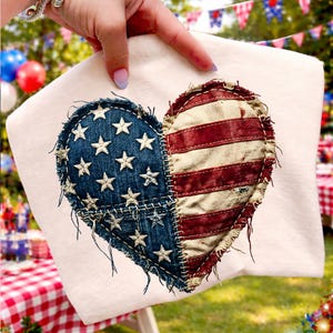 Pode incluir: Um moletom creme com um design de coração. O coração é dividido em duas metades: uma com fundo de ganga azul e estrelas brancas, e a outra com listras vermelhas e brancas, que se assemelham à bandeira americana.