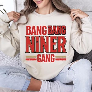 Puede incluir: Sudadera color crema con el texto "BANG BANG NINER GANG" en letras rojas y doradas. La sudadera tiene cuello redondo y mangas largas. El diseño presenta un estilo vintage desgastado.