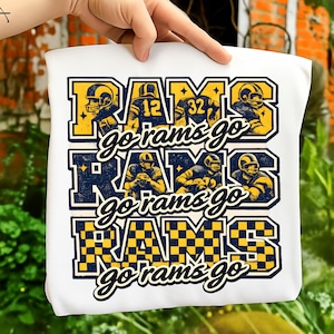 Puede incluir: Camiseta blanca con un diseño azul y amarillo que dice "RAMS go rams go". El diseño incluye jugadores de fútbol americano y los números 12 y 32. La camiseta está sujeta por una mano.