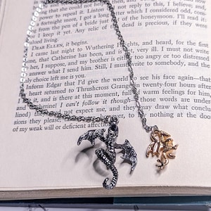 May include: A silver chain necklace with two dragon charms. One charm is a black dragon with wings spread and the other is a gold dragon with wings spread. The necklace is laying on a book with text.