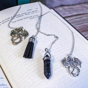 May include: Four silver-toned metal bookmarks with chain attachments rest on an open book. Two feature dragon pendants, one has a black tassel, and another a black crystal pendant. The book is open to a page of printed text.