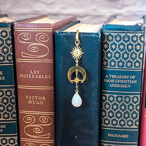 May include: Gold bookmark featuring a crescent moon, star, and a teardrop-shaped white stone pendant. The bookmark is positioned between the spines of three books.