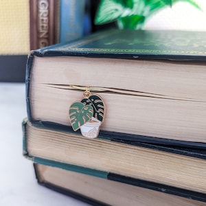May include: Gold bookmark with a green potted plant design. The bookmark is attached to a book with a black spine.