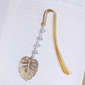 Puede incluir: Un marcapáginas de metal dorado con un dije de hoja y cuentas de cristal transparente. El marcapáginas está sobre las páginas de un libro. El texto "To My Mother" es visible.