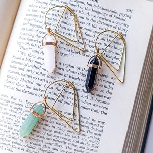 May include: Three gemstone earrings with gold-colored wire frames rest on an open book. One earring features a white crystal, another a black crystal, and the third a light green crystal. The book's pages are aged.