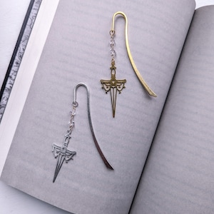Puede incluir: Dos marcapáginas metálicos con forma de espada descansan sobre las páginas abiertas de un libro. Un marcapáginas es de color dorado con un colgante de espada decorativo y cuentas transparentes. El otro es plateado con un colgante similar y cuentas transparentes.