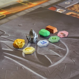 May include: A selection of colorful game pieces, including gold, green, pink, and blue shapes, arranged on a gray game board. The pieces vary in shape and size, with some resembling gems and others rectangular blocks. The board has a textured surface.