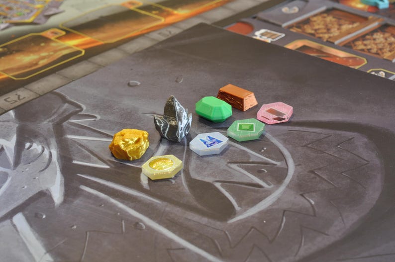 May include: A selection of colorful game pieces, including gold, green, pink, and blue shapes, arranged on a gray game board. The pieces vary in shape and size, with some resembling gems and others rectangular blocks. The board has a textured surface.