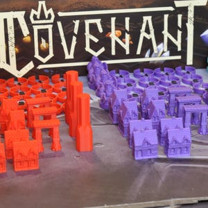 May include: A selection of colorful miniature game pieces, including red buildings, purple structures, and teal towers. The word "COVENANT" is displayed in white above the pieces. The pieces are arranged on a gray surface.