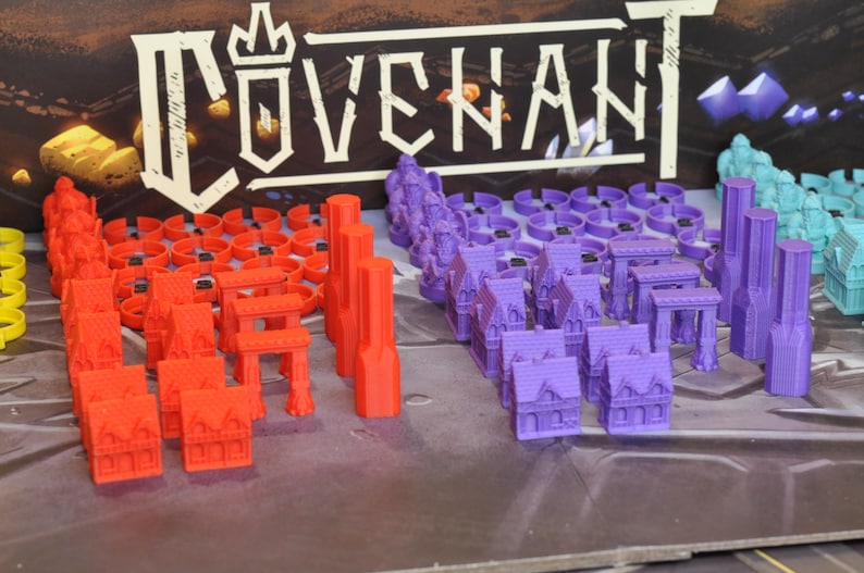 May include: A selection of colorful miniature game pieces, including red buildings, purple structures, and teal towers. The word "COVENANT" is displayed in white above the pieces. The pieces are arranged on a gray surface.