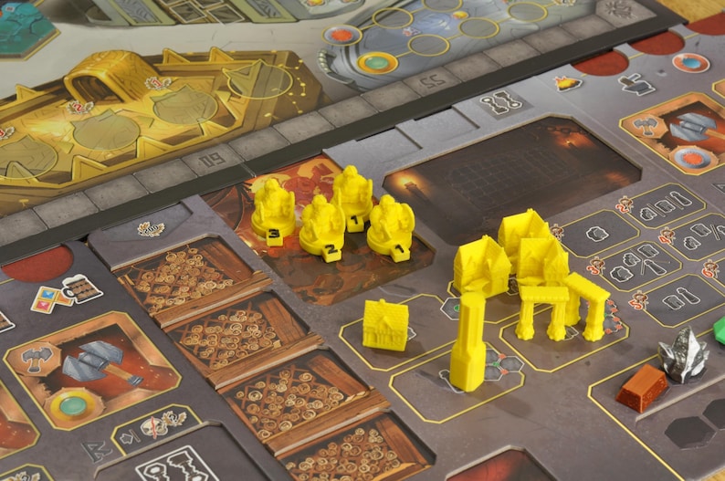 May include: A board game with a detailed game board featuring various sections and yellow game pieces. The board has a metallic look with brown and gray tones. Yellow game pieces include figures, chairs, and other structures. The game appears to be a strategy or resource management game.