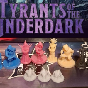 Może przedstawiać: Zestaw figurek do gry planszowej "Tyrants of the Underdark". Figurki są wykonane z plastiku i pomalowane w różnych kolorach, w tym niebieskim, różowym, beżowym i czarnym. Figurki stoją na okrągłych podstawach.