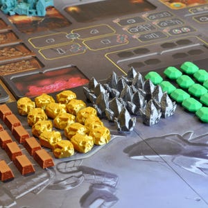 May include: A board game setup with various game pieces. There are gold-colored rock-like pieces, silver-colored crystal-like pieces, and green gem-shaped pieces. Also, there are brown rectangular pieces and a game board.