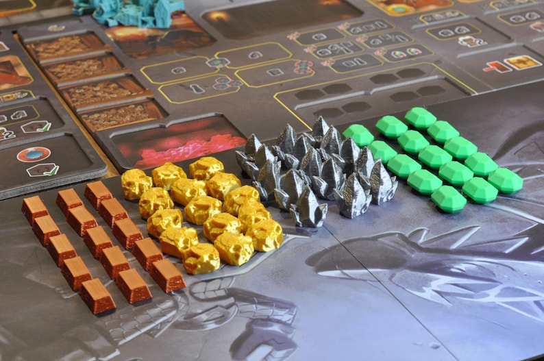 May include: A board game setup with various game pieces. There are gold-colored rock-like pieces, silver-colored crystal-like pieces, and green gem-shaped pieces. Also, there are brown rectangular pieces and a game board.