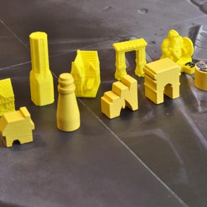 May include: A collection of yellow game pieces, including miniature houses, a lighthouse, archways, and a statue. The pieces are arranged on a gray surface, suggesting a board game or tabletop activity. The pieces are made of wood or plastic.