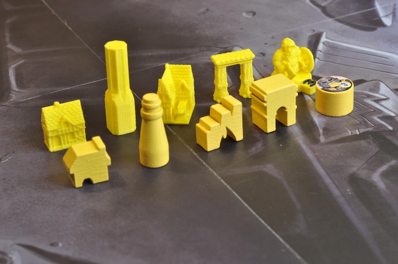 May include: A collection of yellow game pieces, including miniature houses, a lighthouse, archways, and a statue. The pieces are arranged on a gray surface, suggesting a board game or tabletop activity. The pieces are made of wood or plastic.