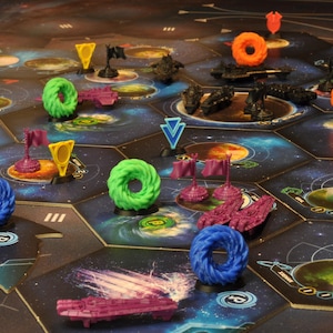 Op de afbeelding: Een bordspel met een ruimtethema. Het spelbord bestaat uit zeshoekige tegels met planeten en sterrenstelsels. Er zijn verschillende spelstukken, waaronder schepen, vlaggen en ringvormige markeringen in blauw, groen, oranje en paars.