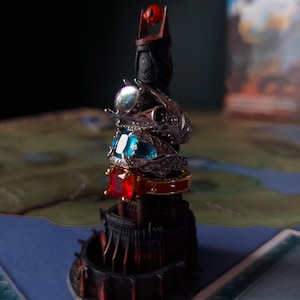 以下が含まれることがあります： 赤い目をつけた黒い塔が、カラフルな宝石がついた4つの指輪に囲まれている。指輪は互いに積み重ねられており、銀と金のようだ。